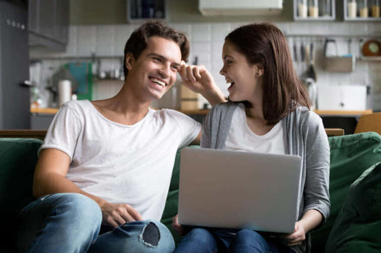 幸せなカップルが、台所のソファでノートパソコンと一緒に笑い合い、笑顔の若い女の子と男性が家で楽しみながらコンピュータを使ってソファで話している