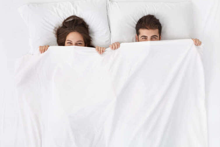 可笑しい夫婦が寝そべって白い毛布の下に隠れ、喜びに満ちた目でカメラを見つめている