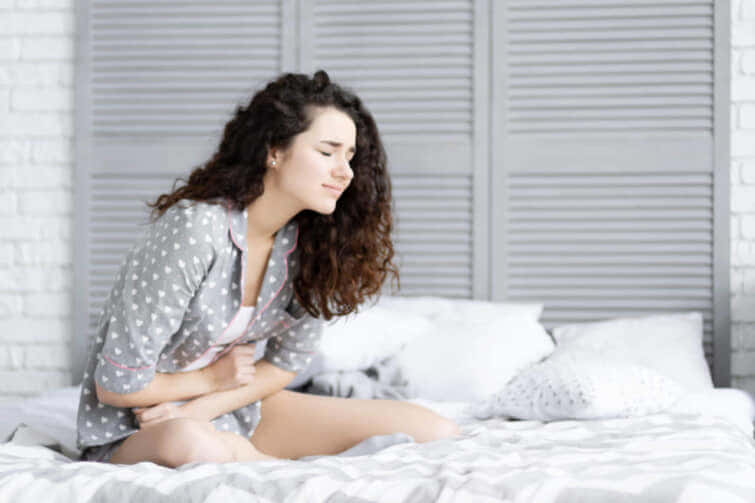 巻き毛と灰色のパジャマを持つブルネットの女性女性は、ベッドに腰を下ろし、腹痛で寝ている。医療、眠れない月経のコンセプト