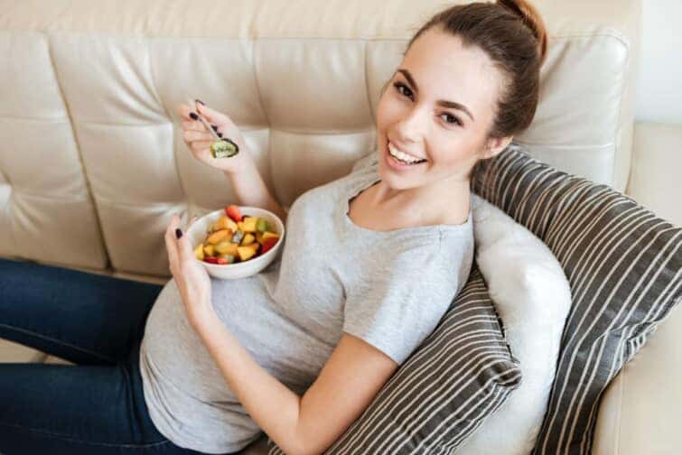 家のソファでフルーツサラダを食べながら座って幸せな妊婦