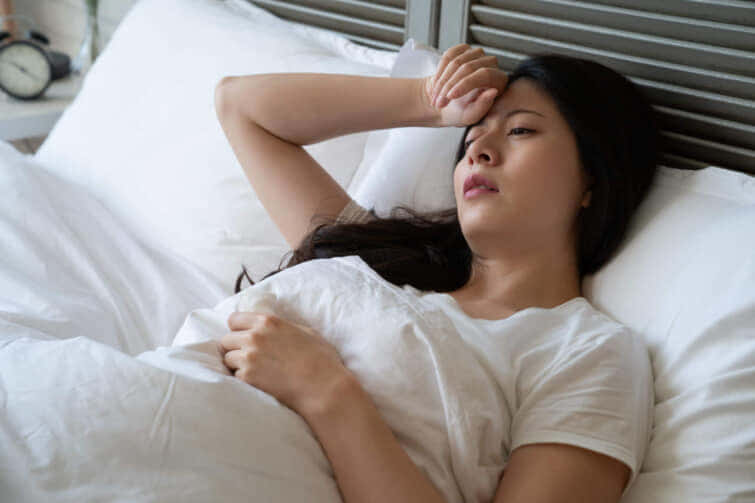 体温を測る手を手にして頭痛を感じるアジア系の病人が、自宅のベッドに寝転がっています。寝室で昼間だけ休む白い暖かい毛布を持つ、女性の体を覆う体。