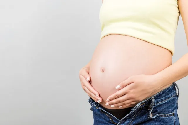 コピー スペースとカラフルな背景で妊娠中の腹のクローズ アップ。 開いたジーンズの将来の母親は、彼女の裸の赤ちゃんの隆起を抱きしめています. 母性の概念