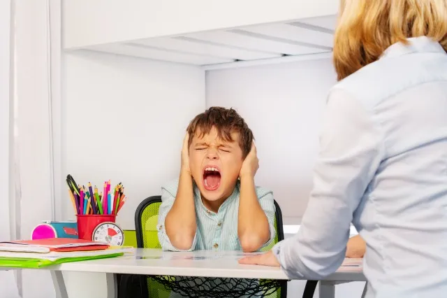自閉症の少年は、教師と一緒にテーブルのそばに座っている ABA 発達療法中に悲鳴を上げ、痛みで耳を閉じる