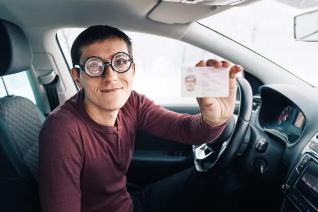 巨大なばかげた眼鏡をかけた面白いオタクとオタクは、彼の運転免許証を示しています。 視力矯正の概念と道路上の愚か者