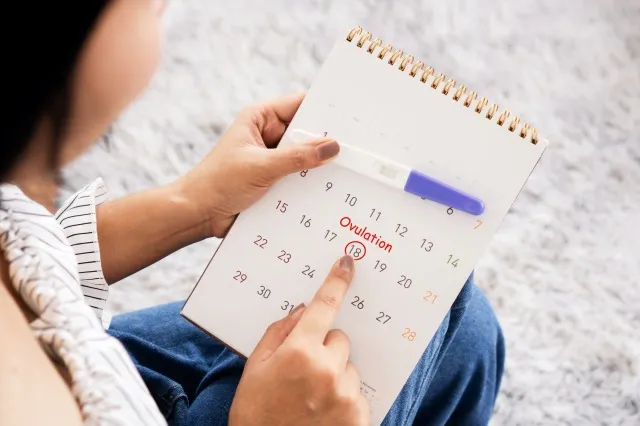 排卵日のための彼女の月経周期の計画をチェックするカレンダーの日付を数えている女性の手妊娠検査を持っている別の手