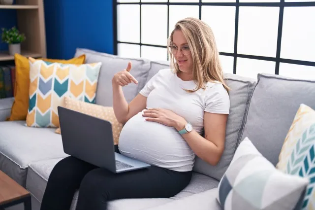 ラップトップを使用して自宅のソファに座っている若い妊婦の手と指で幸せなポインティングを笑顔
