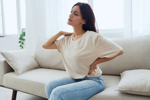 自宅のソファに座っている女性 背中と腰の痛み、突起、30年後の背中の問題