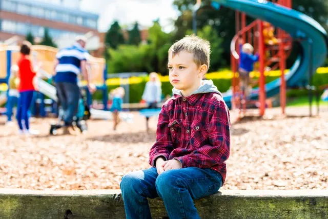 公園に座っているADHD、自閉症、アスペルガー症候群のハンサムな少年は、他の子どもたちと遊ぶことに対して恐怖と不安を感じ、緊張と不安を感じている