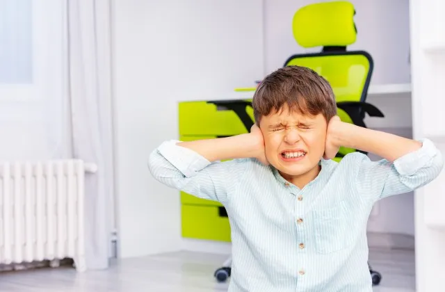 自閉症の学齢で、強い否定的な顔つきの、耳を閉じ、歯を磨く少年が部屋に座っている