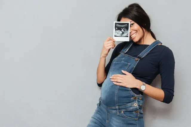 グレイの背景に超音波スキャンを見せながら、立ってポーズをとる妊婦の笑顔の女性の画像
