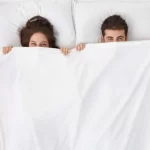 可笑しい夫婦が寝そべって白い毛布の下に隠れ、喜びに満ちた目でカメラを見つめている